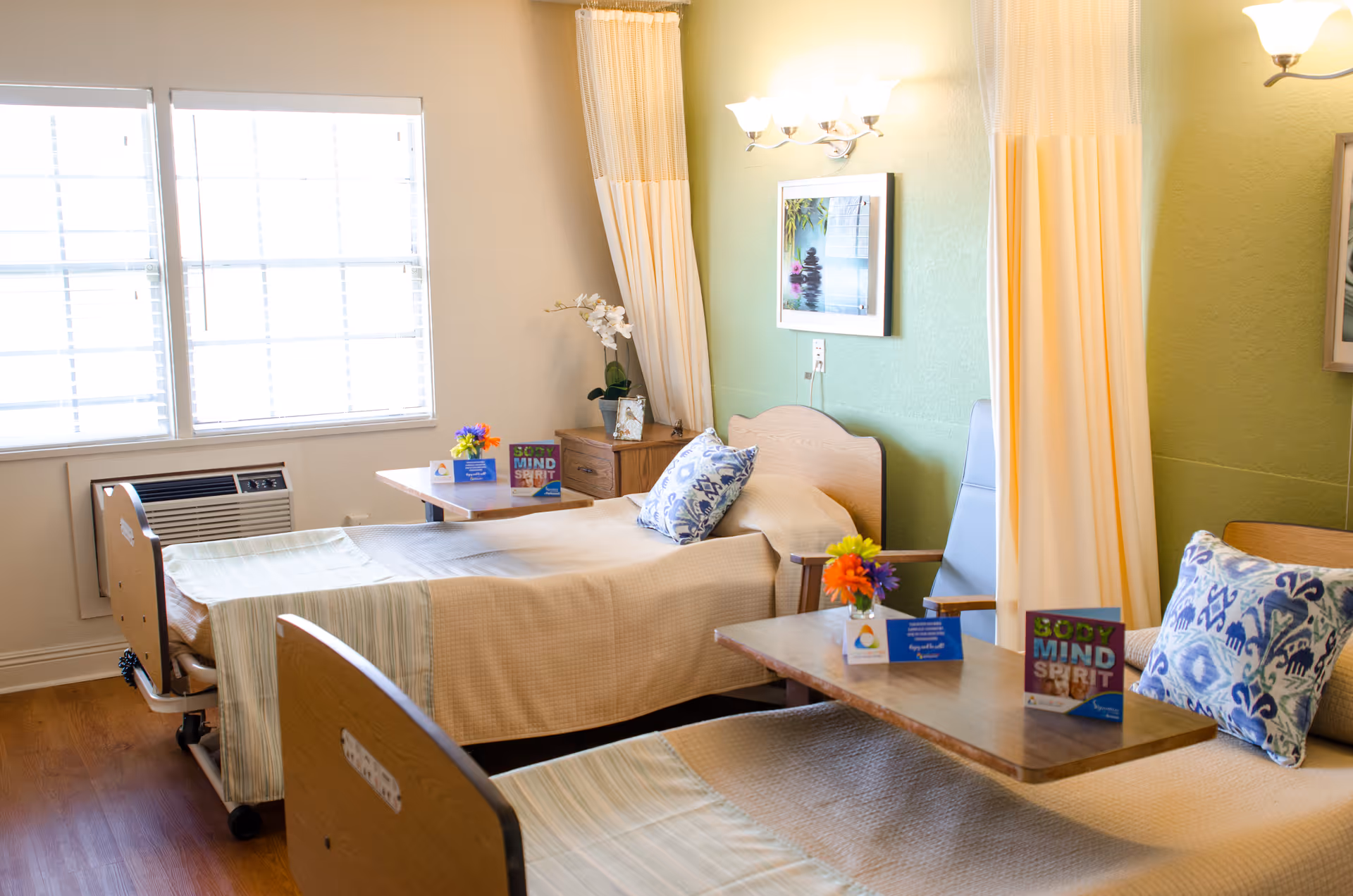 A bright and clean bedroom in a senior living facility featuring two hospital-style beds with beige bedding and patterned pillows. Each bed has a wooden bedside table with a small vase of colorful flowers and informational cards. The room has a large window with blinds, light green walls, a framed picture, and privacy curtains between the beds.