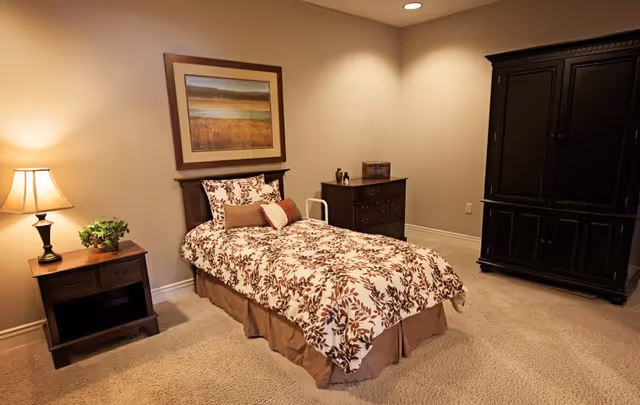 A cozy bedroom with a single bed covered in a floral patterned bedspread. Next to the bed is a wooden nightstand with a lamp and a small plant. On the wall above the bed hangs a framed landscape painting. Across from the bed is a dark wooden dresser and a large matching armoire. The room has beige walls and carpeted floor with recessed lighting in the ceiling.