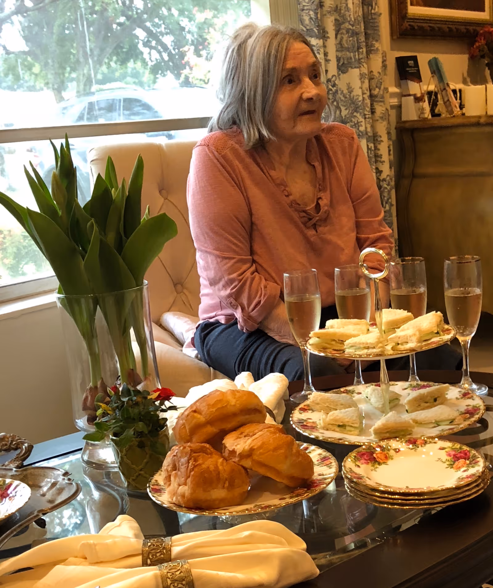 An elderly woman with gray hair wearing a pink blouse sits on a beige armchair near a window. In front of her is a glass table with a vase of green tulips, a small potted plant, a plate of croissants, a tiered tray with finger sandwiches, several glasses of a light beverage, and stacked floral plates with napkins.