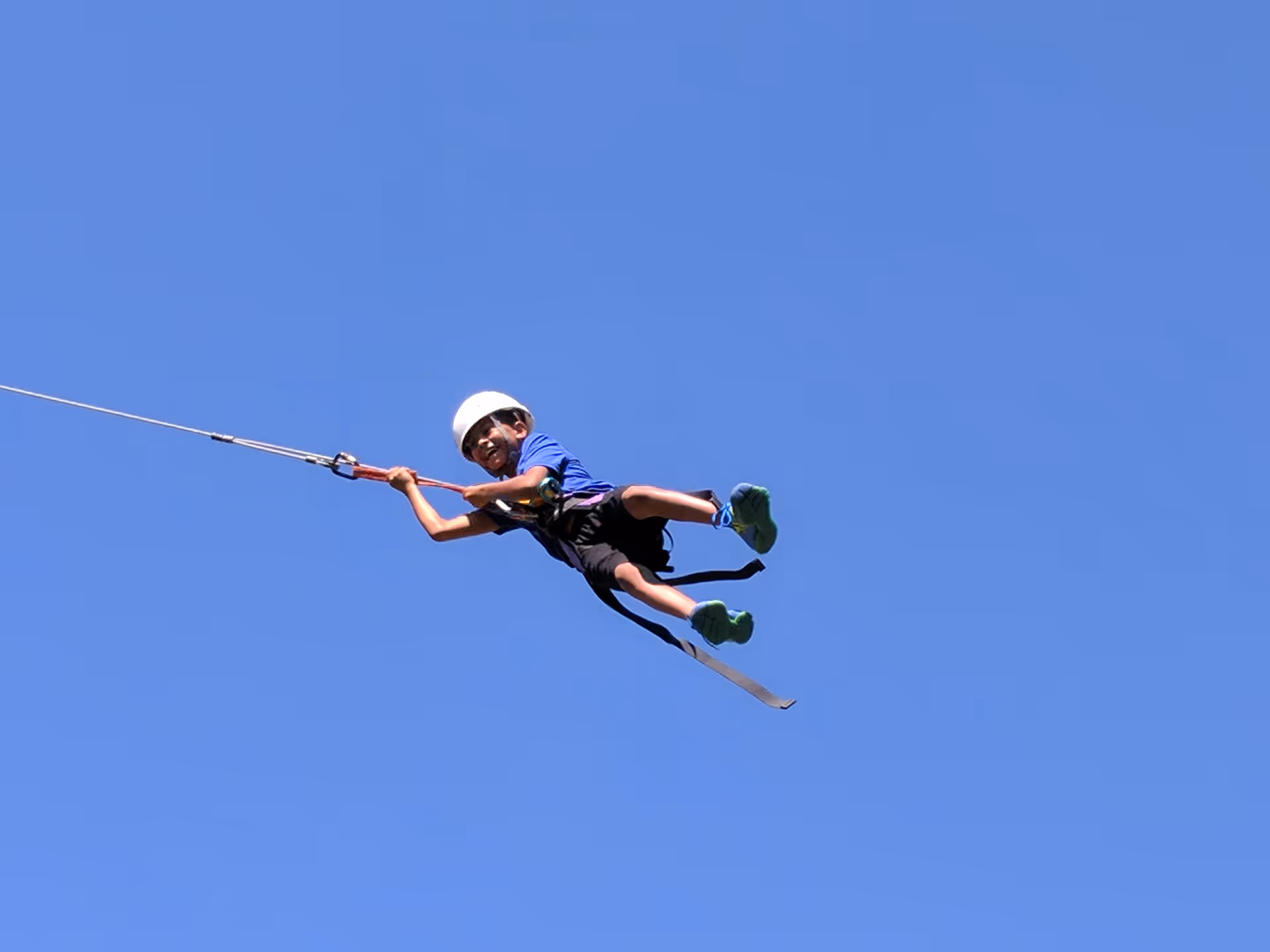 Person wearing a helmet zip-lining through a clear blue sky.