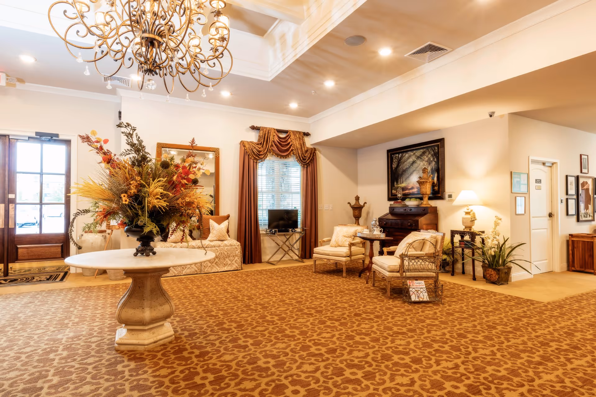 Warm, well-lit lobby/common sitting area with a chandelier, a central round table holding a large floral arrangement, and upholstered seating along the walls.