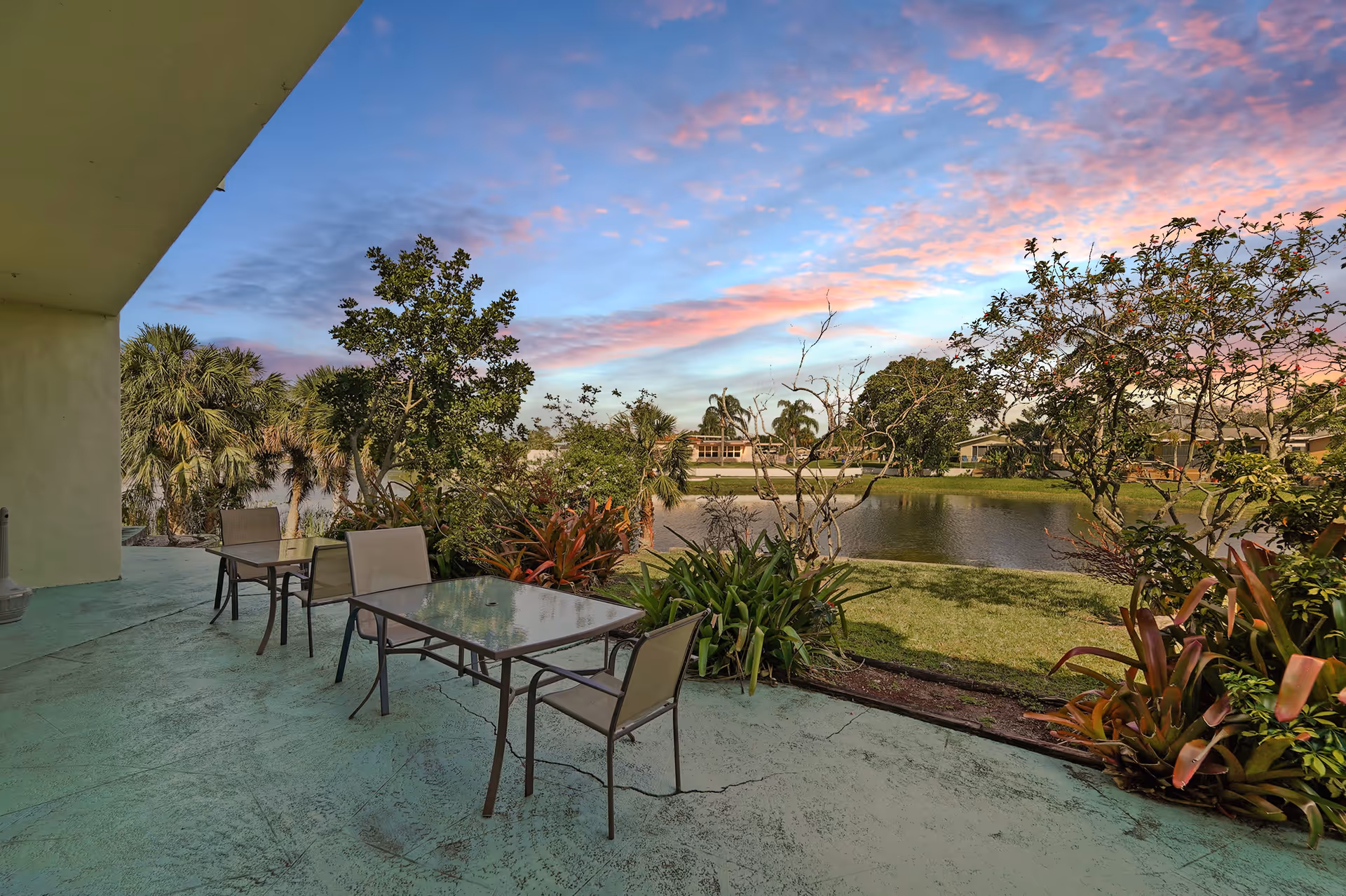 Outdoor patio with glass-top tables and chairs overlooking a landscaped pond and colorful sunset sky.