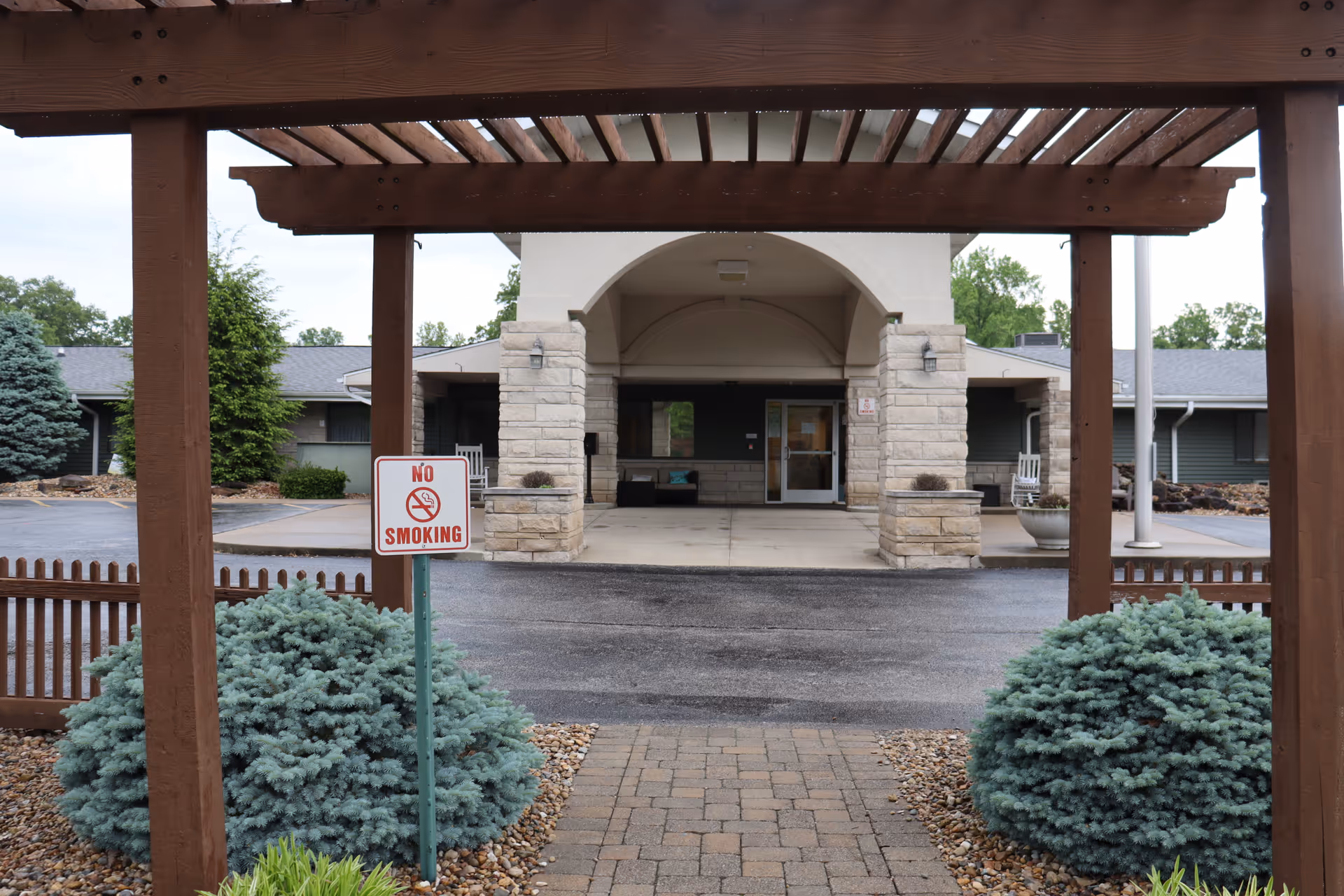 View of the entrance to a building with a covered walkway supported by stone pillars. There is a wooden pergola structure in the foreground and a 'No Smoking' sign posted near some green shrubs. The building has a dark green exterior with a gray roof and a paved driveway leading to the entrance.