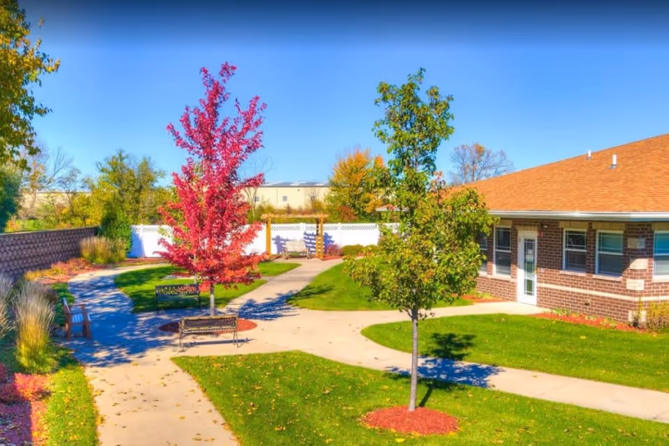 Outdoor garden area at River Valley Place of Ottumwa with paved walkways, green grass, benches, and trees including one with bright red leaves. A brick building with a brown roof is visible on the right side under a clear blue sky.