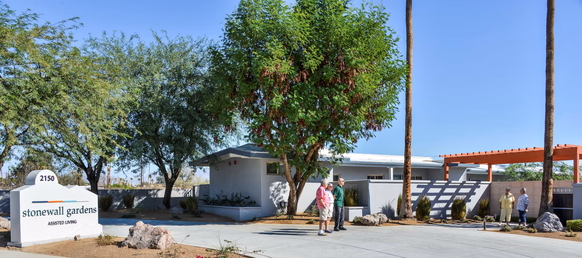 Front entrance of Stonewall Gardens assisted living with a sign, low modern building, trees, a pergola, and several people standing outside.