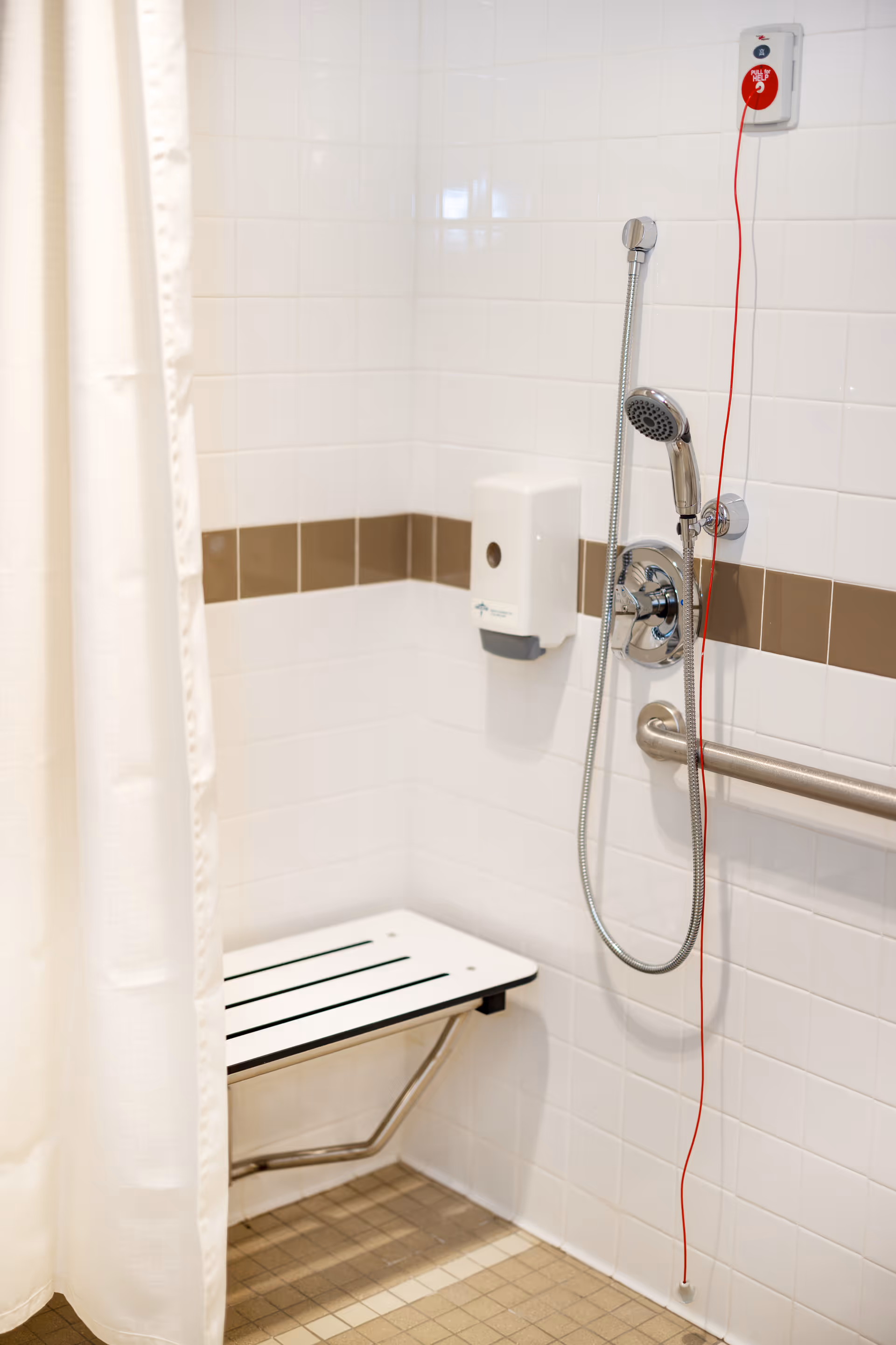 Accessible shower with bench, handheld showerhead, white tiles, and curtain in a senior living bathroom.