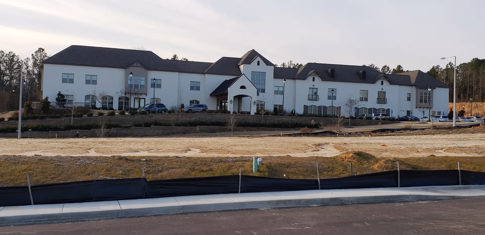 White senior living facility exterior with arched entry, cars in lot, construction fencing in front.