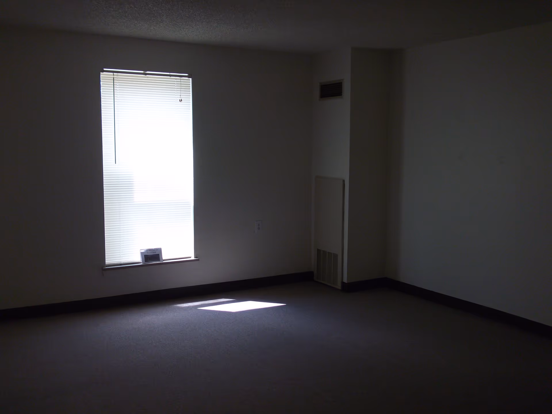 Empty dimly lit carpeted room with a single window with blinds and a wall vent.