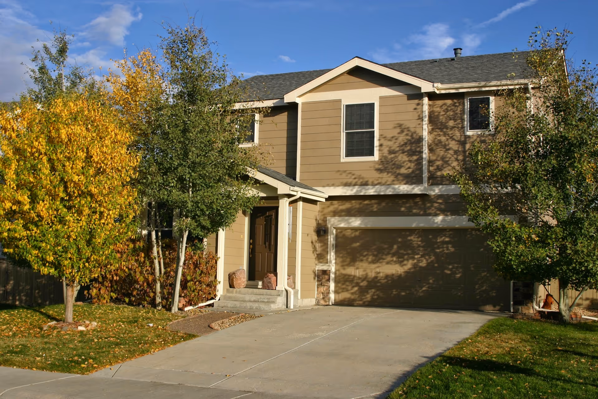 Two-story beige house with attached garage and autumn trees along a concrete driveway.