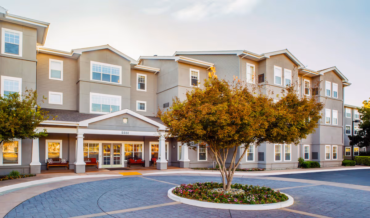 Three-story senior living building with a covered entrance, benches, and a landscaped circular driveway with a tree.