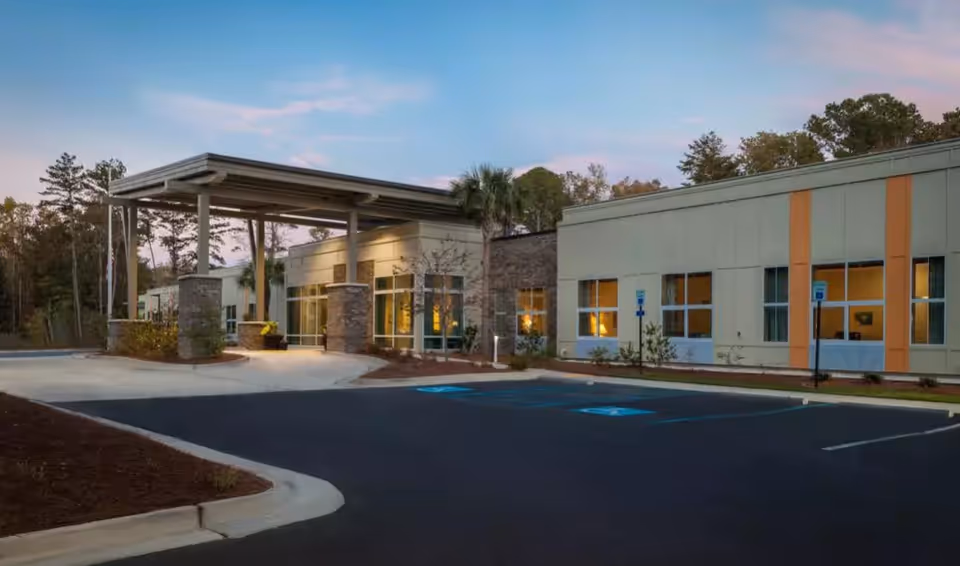 Exterior view of Ashley Gardens Assisted Living & Memory Care building at dusk, showing the entrance with a covered drop-off area, parking spaces including handicapped spots, and surrounding trees.