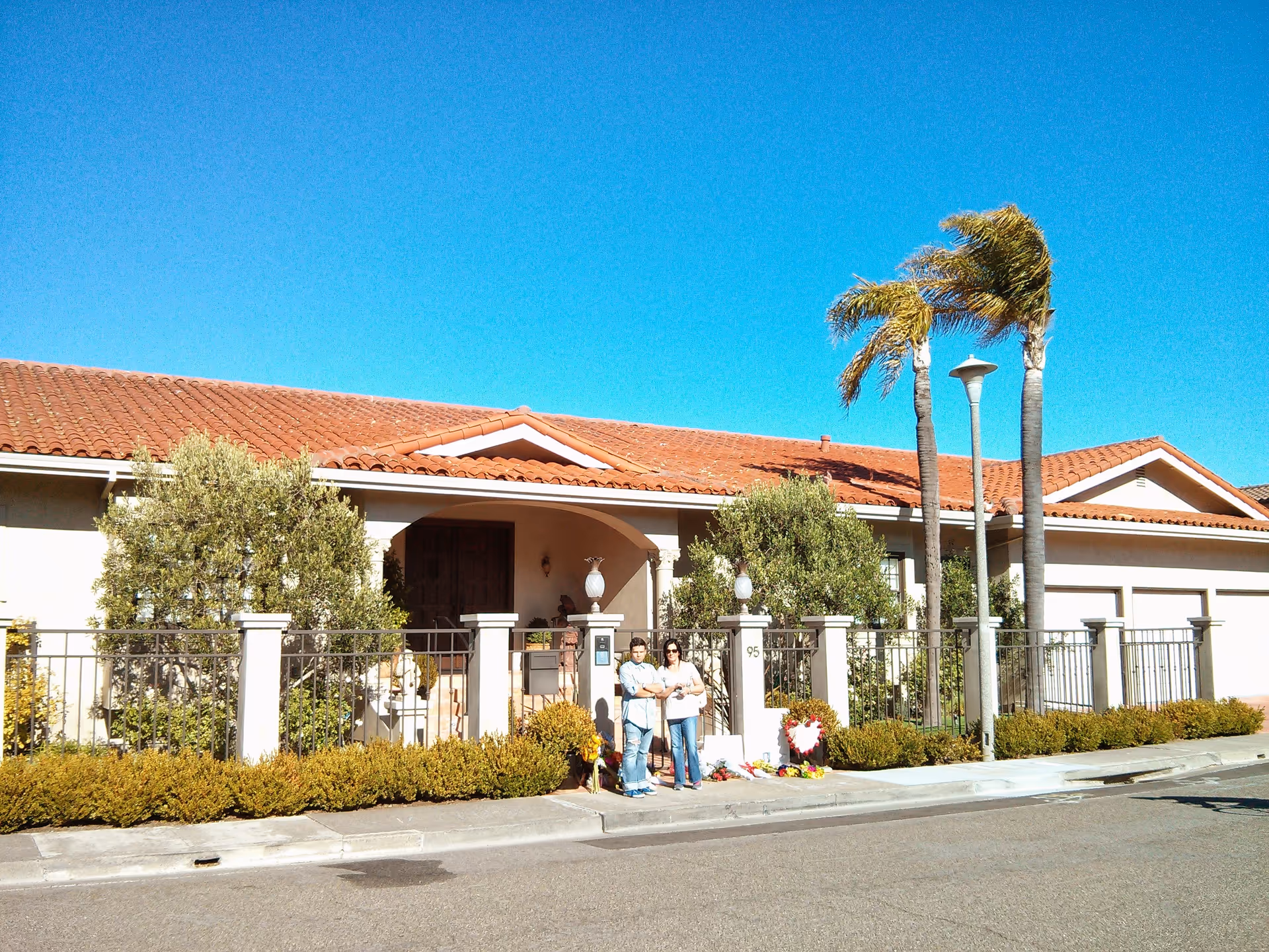 Single-story senior living facility with red tile roof, fence, plants, and two people at entrance.