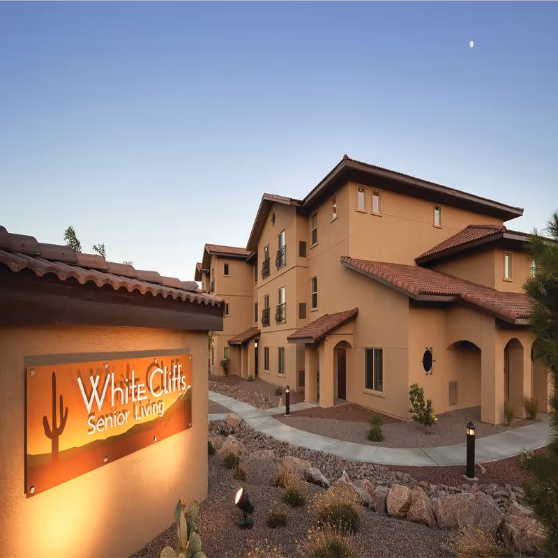Exterior view of White Cliffs Senior Living facility during dusk, showing a multi-story building with tan stucco walls and red tile roofing, surrounded by desert landscaping with rocks and plants. A lit sign with the facility name and a cactus graphic is visible in the foreground.