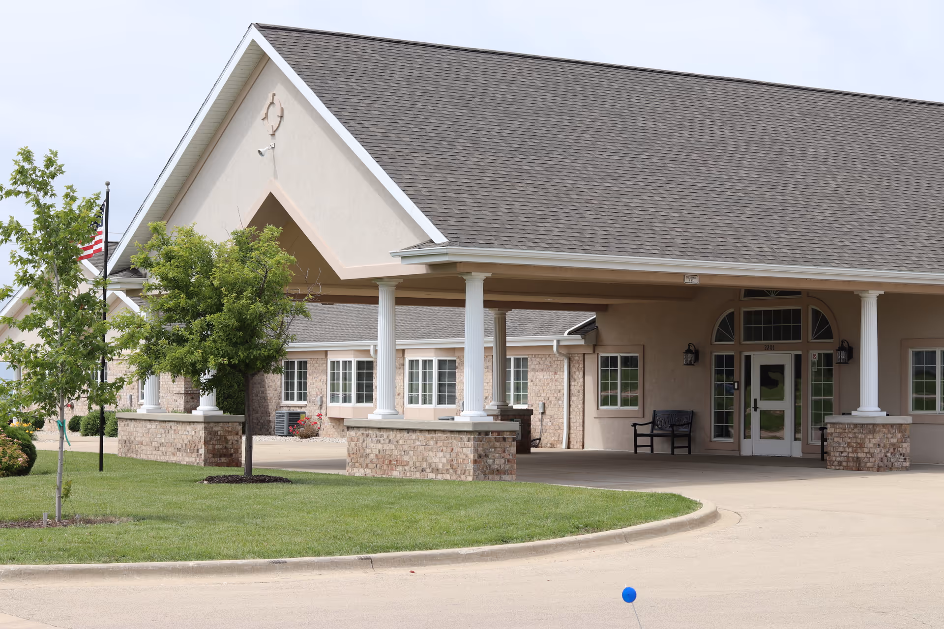 Exterior view of Liberty Village of Rochelle building entrance with a covered driveway supported by white columns, brick and beige walls, several windows, a bench near the door, and a small tree and American flag on the lawn.