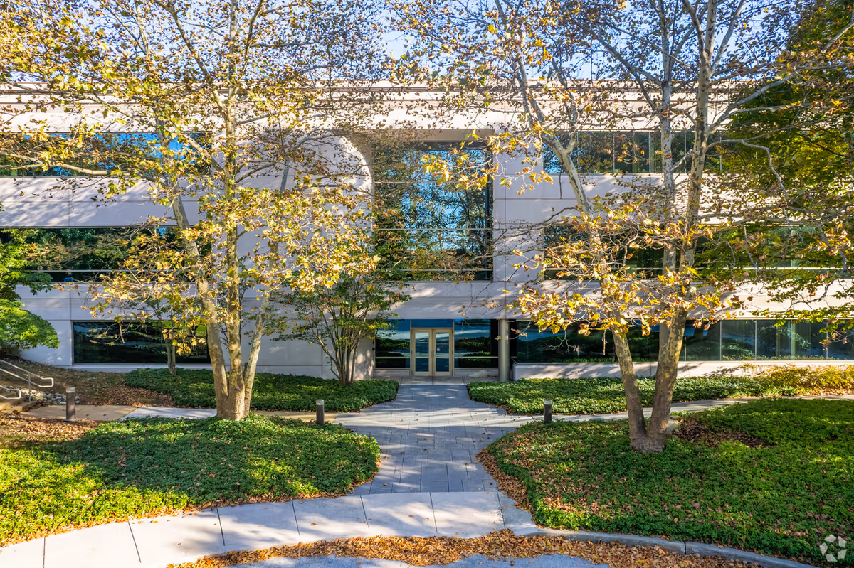 Modern low-rise building exterior with glass entrance, walkway, and landscaped trees.
