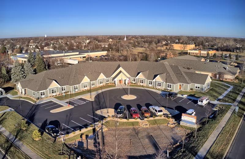 Aerial view of a single-story U-shaped senior living building with a circular driveway, parked cars, and surrounding neighborhood.