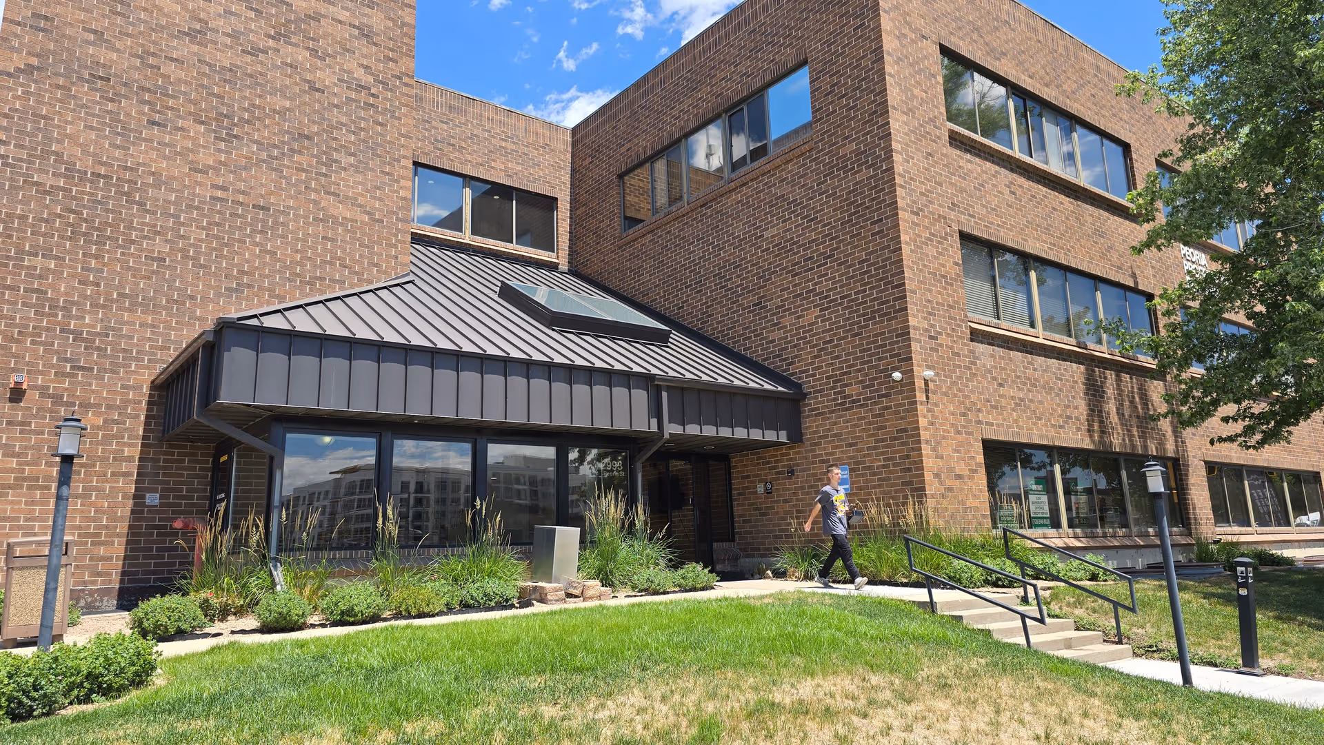 Brick senior living facility exterior with glass entrance, awning, landscaping, and a person walking outside.