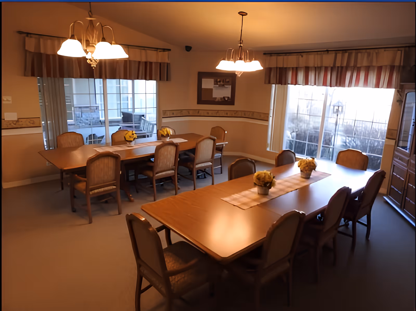 A dining room with two wooden tables, each surrounded by chairs. The tables have beige runners and small flower arrangements. Large windows with striped valances let in natural light. Two ceiling light fixtures hang above the tables, and a mirror is mounted on the wall.