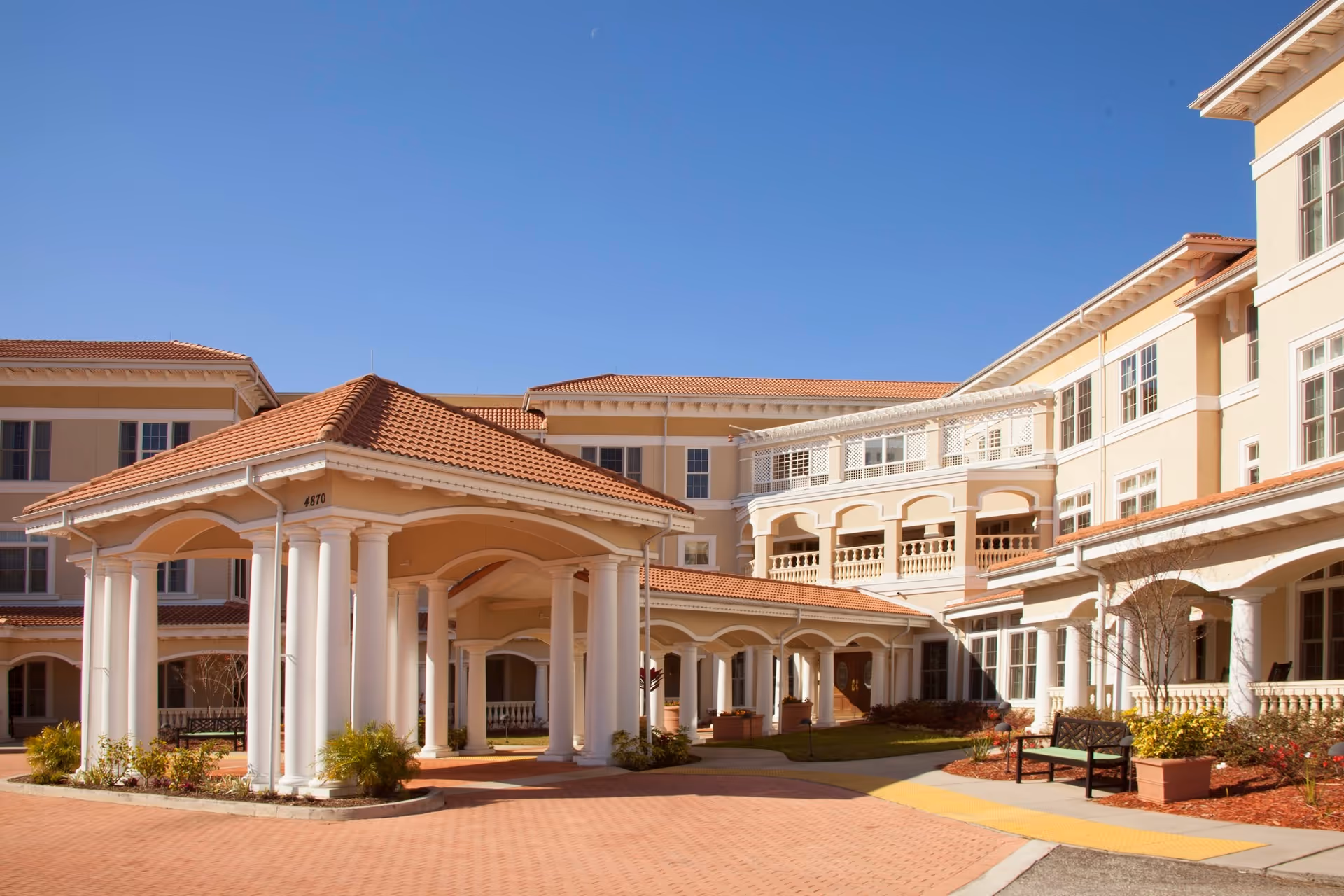 Exterior view of a large senior living facility building with beige walls and red tile roofs under a clear blue sky. The entrance features a covered driveway supported by white columns, with benches and landscaped plants nearby.