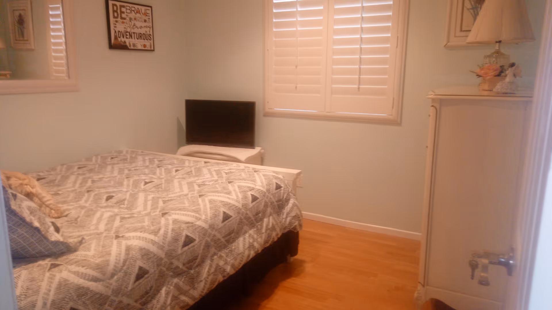 A small bedroom with a bed covered in a patterned gray and white comforter, a pillow, a small TV on a white stand, a window with white shutters, a white dresser with a lamp and decorative items, and a framed inspirational quote on the wall.