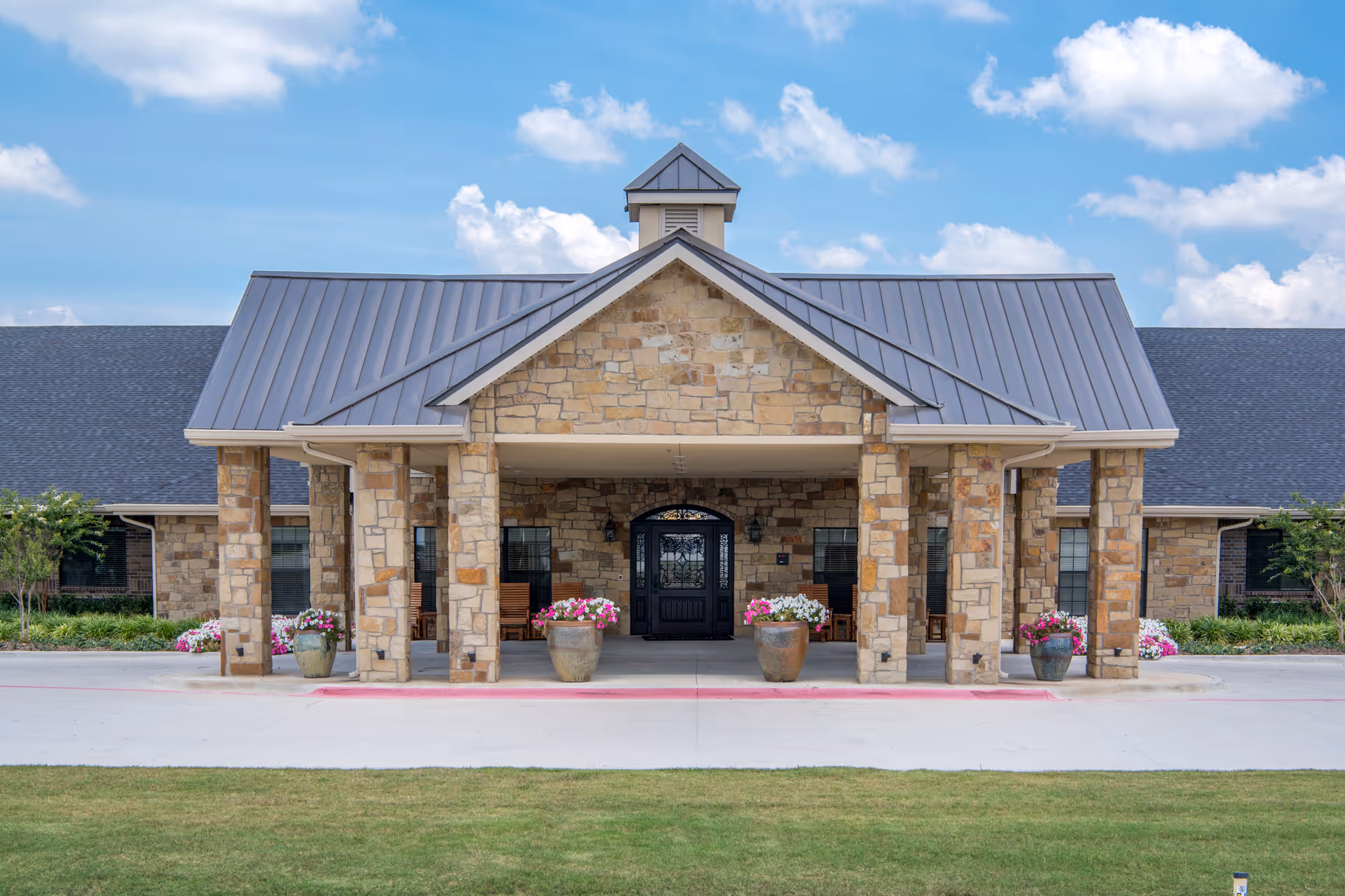 Front exterior view of Lexington Medical Lodge featuring a stone facade with a covered entrance supported by stone pillars, potted flowers, and a black double door under a blue sky with scattered clouds.