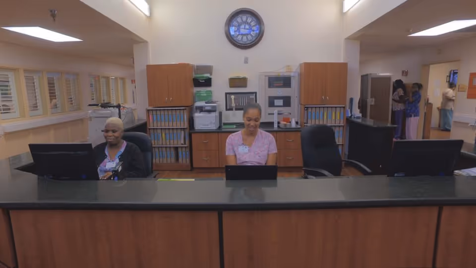 Reception desk in a nursing facility lobby with two staff seated behind a long counter and cabinets with files in the background.