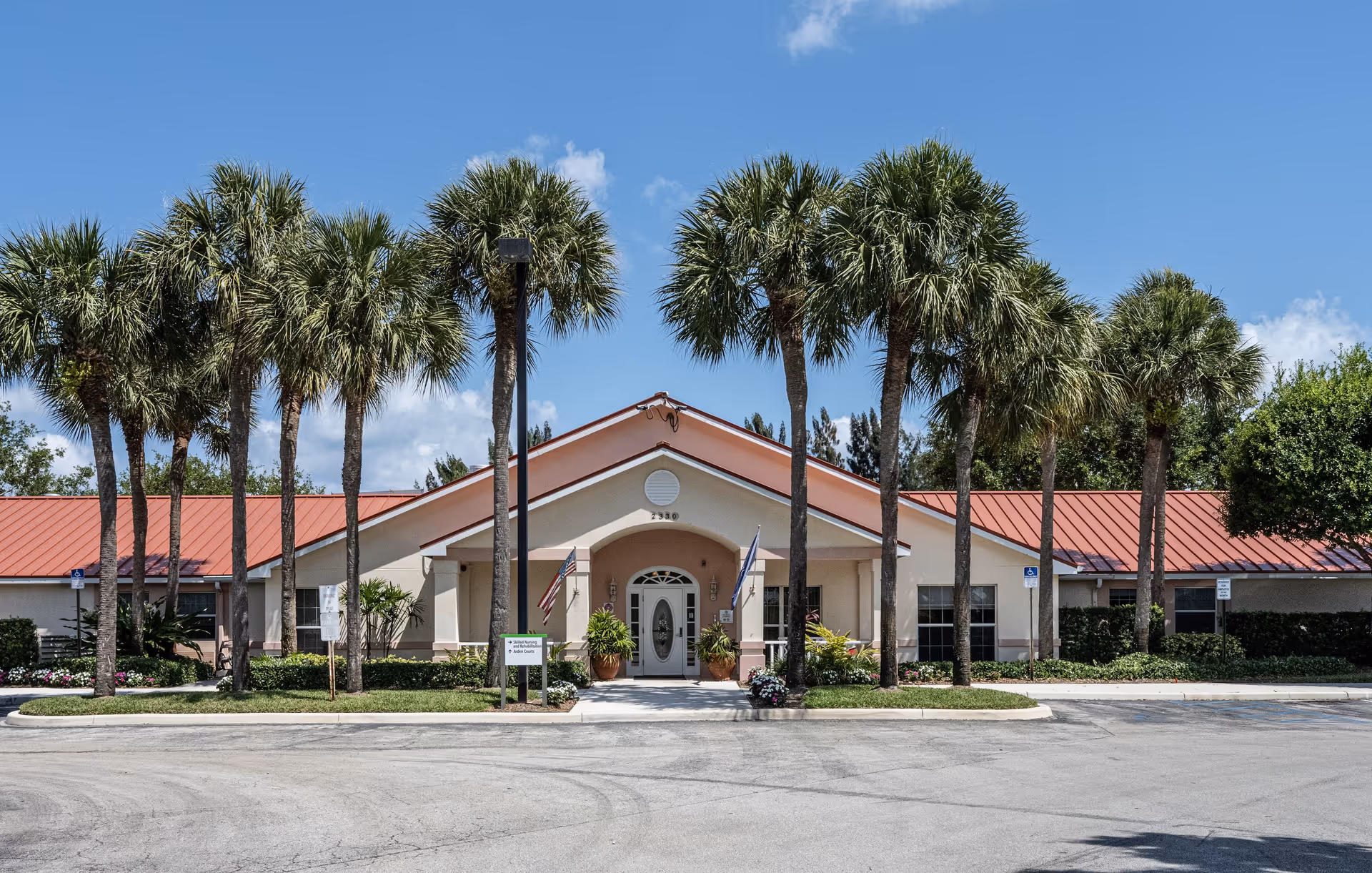 Exterior view of a single-story building with a red roof and beige walls, surrounded by tall palm trees and landscaping, under a clear blue sky.