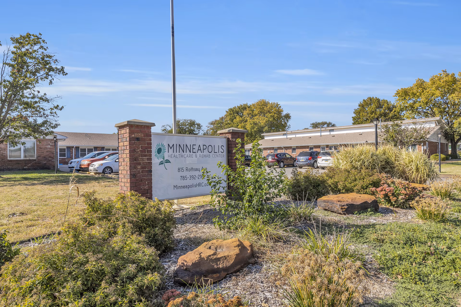 Exterior view of Minneapolis Healthcare & Rehab Center with a large sign in front displaying the facility name, address, phone number, and website. The building is a single-story brick structure with several parked cars visible in the parking lot. There are trees and landscaped bushes around the sign and building under a clear blue sky.