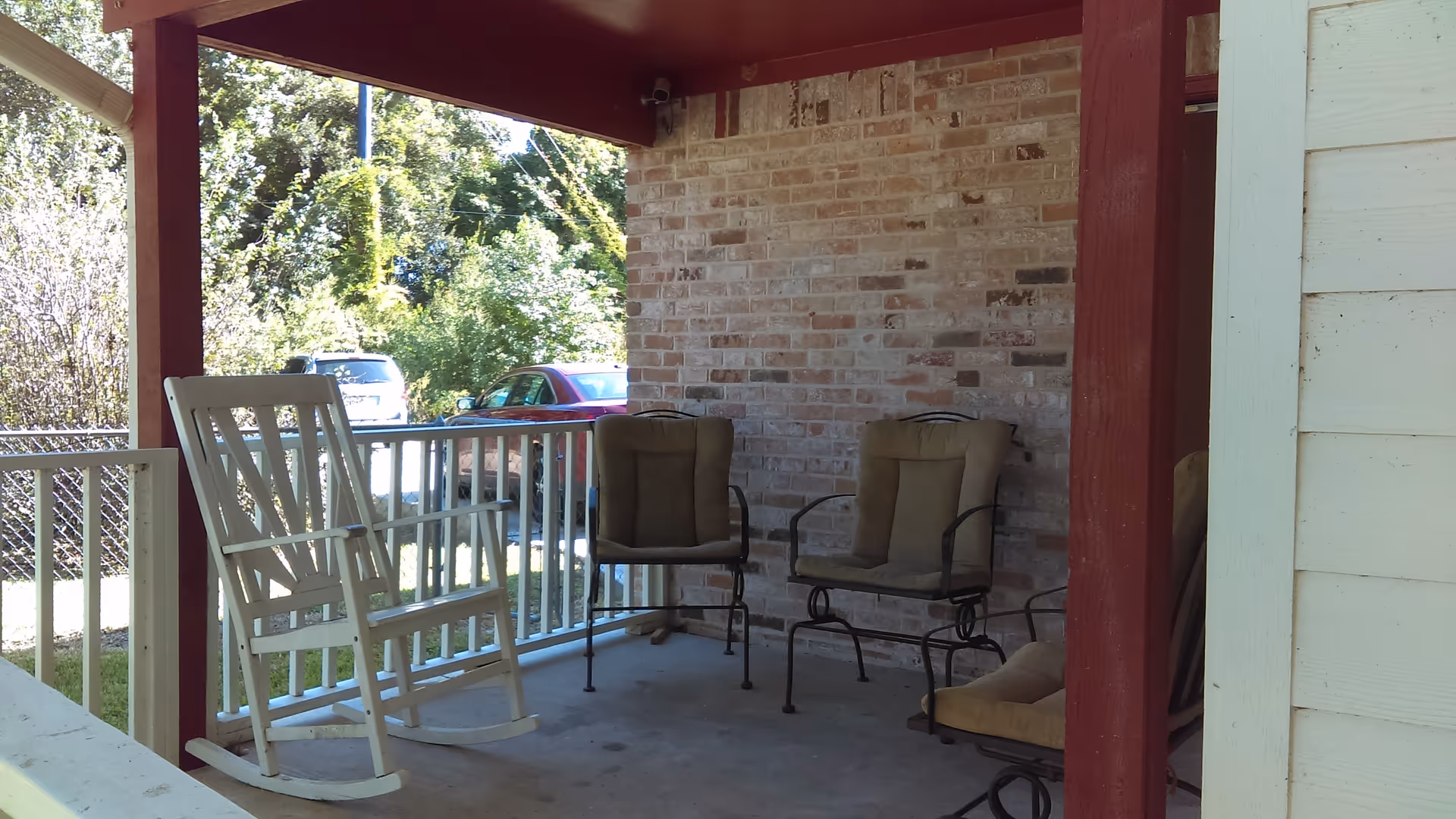 Covered front porch with a white rocking chair and several cushioned metal chairs beside a brick wall.