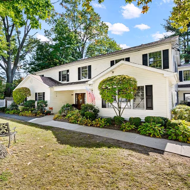 Exterior view of a two-story white building with black shutters, surrounded by green trees and shrubs. A concrete walkway leads to the entrance, which has a small covered porch with an American flag hanging. The sky is blue with some clouds.