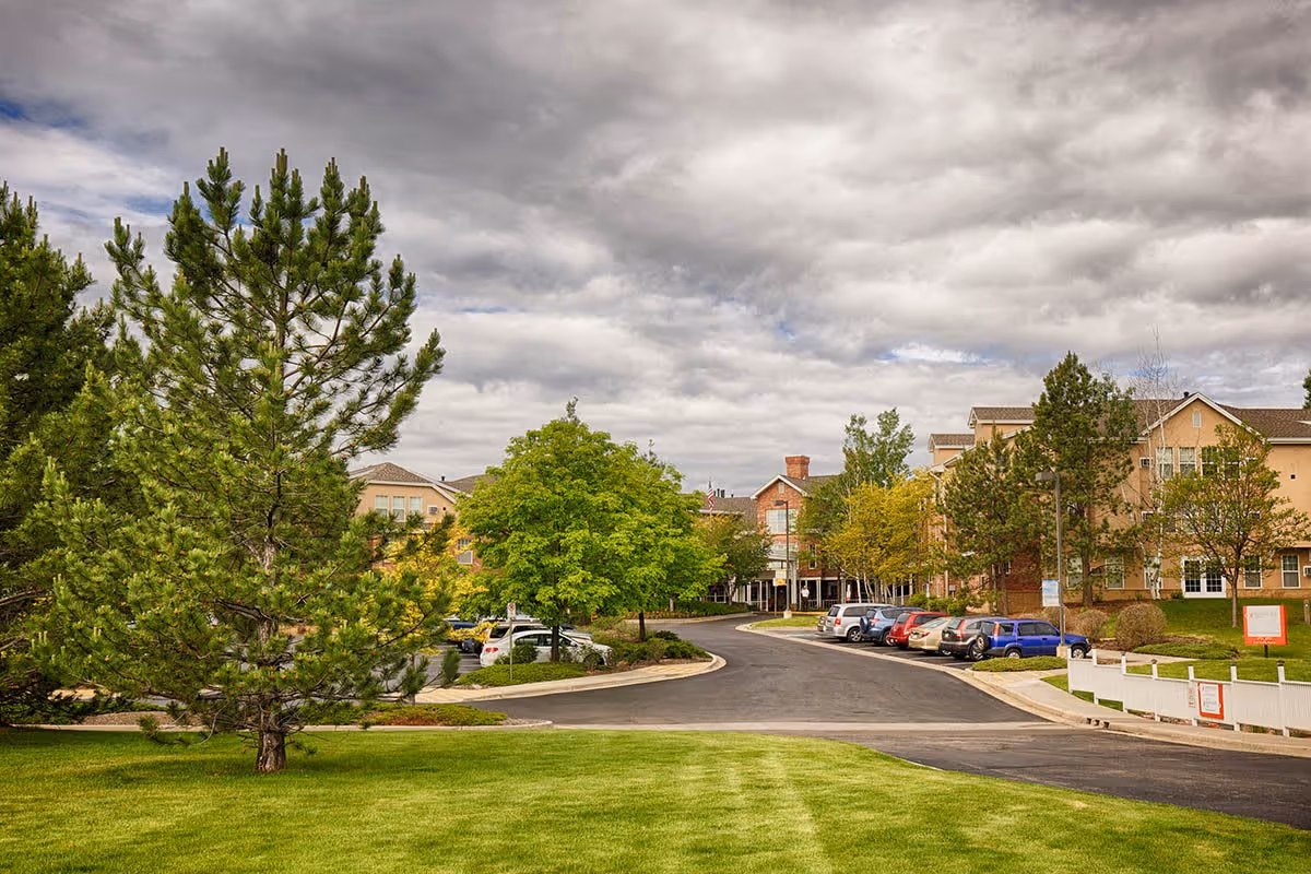 View of the Someren Glen Retirement Community showing a paved driveway lined with parked cars, surrounded by green grass, trees, and multi-story residential buildings under a cloudy sky.