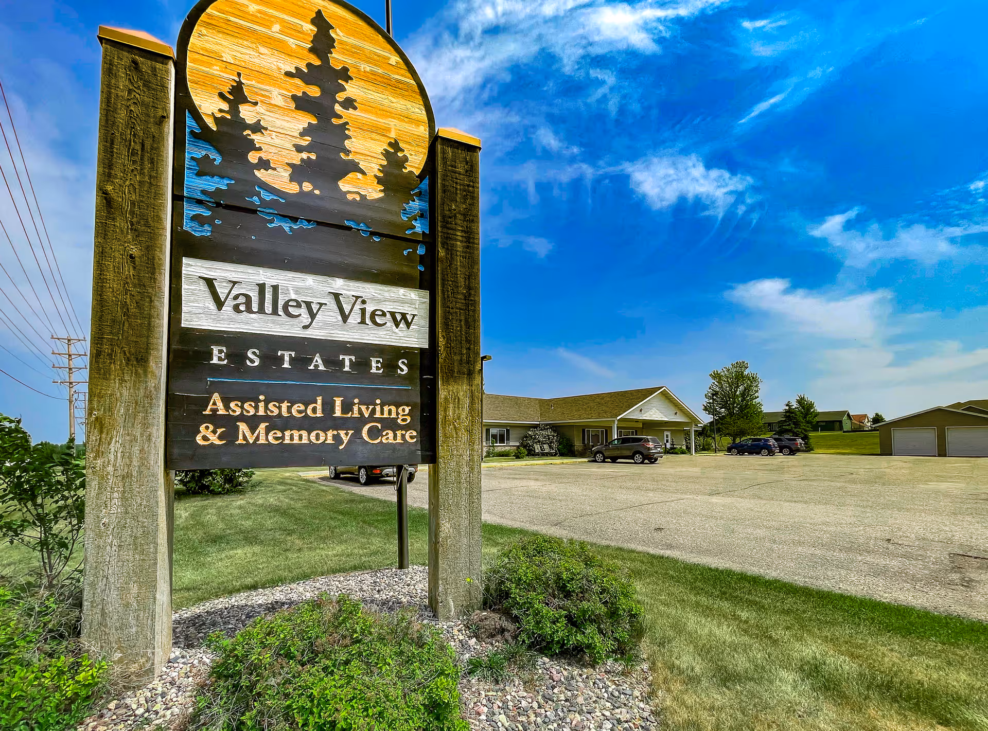 Outdoor view of Valley View Estates Assisted Living & Memory Care sign with a building, parking lot, cars, grass, and blue sky in the background.