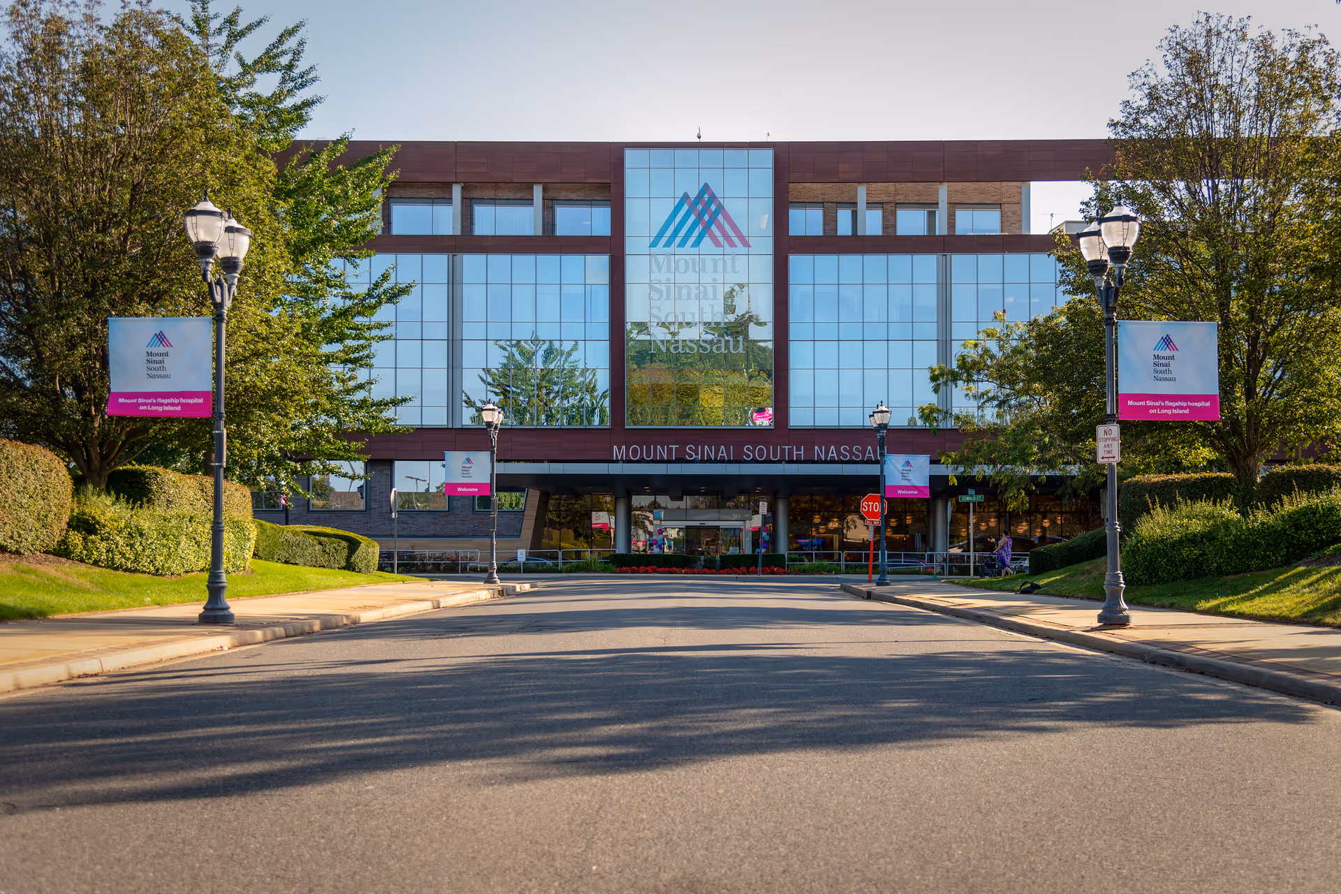 Bright hospital building front with glass facade, banners, trees, and a wide driveway.