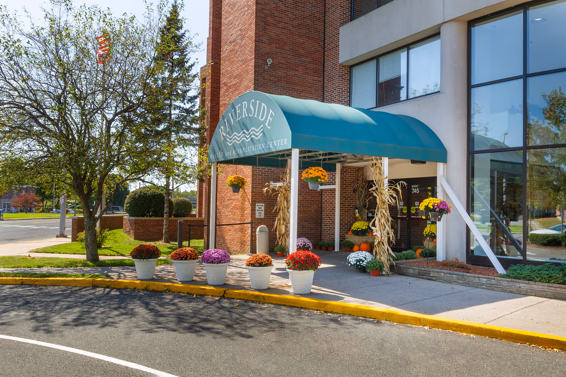 Entrance of Riverside Health and Rehabilitation Center with a green awning displaying the facility name. The entrance is decorated with colorful potted flowers and autumn-themed decorations including pumpkins and cornstalks. The building has large windows and a brick exterior, with a tree and a flagpole visible nearby.