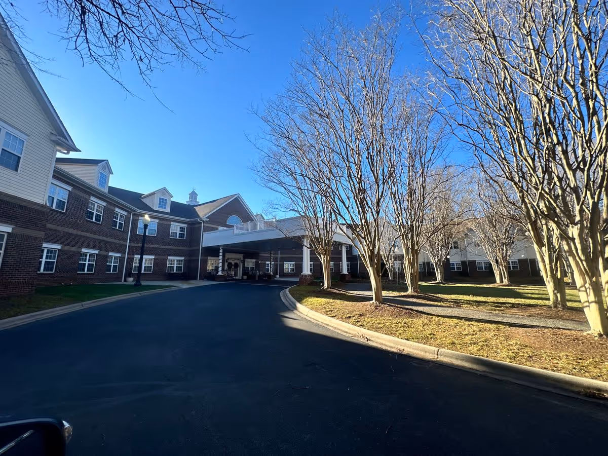 Front entrance of a brick senior living building with a covered porte-cochère, circular driveway and leafless trees under a clear blue sky.