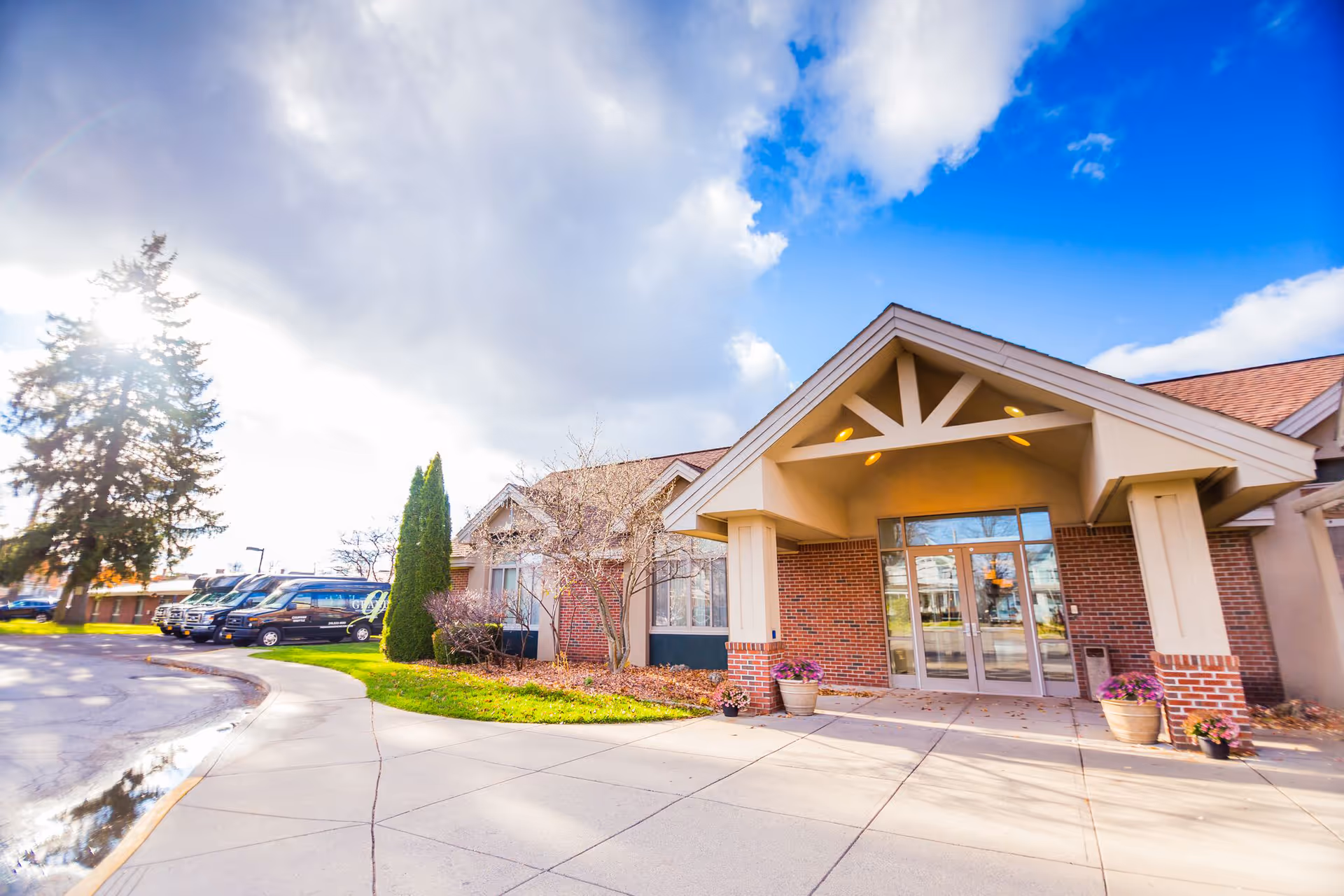 Front entrance of a brick senior living facility with a covered entry and parked cars nearby.