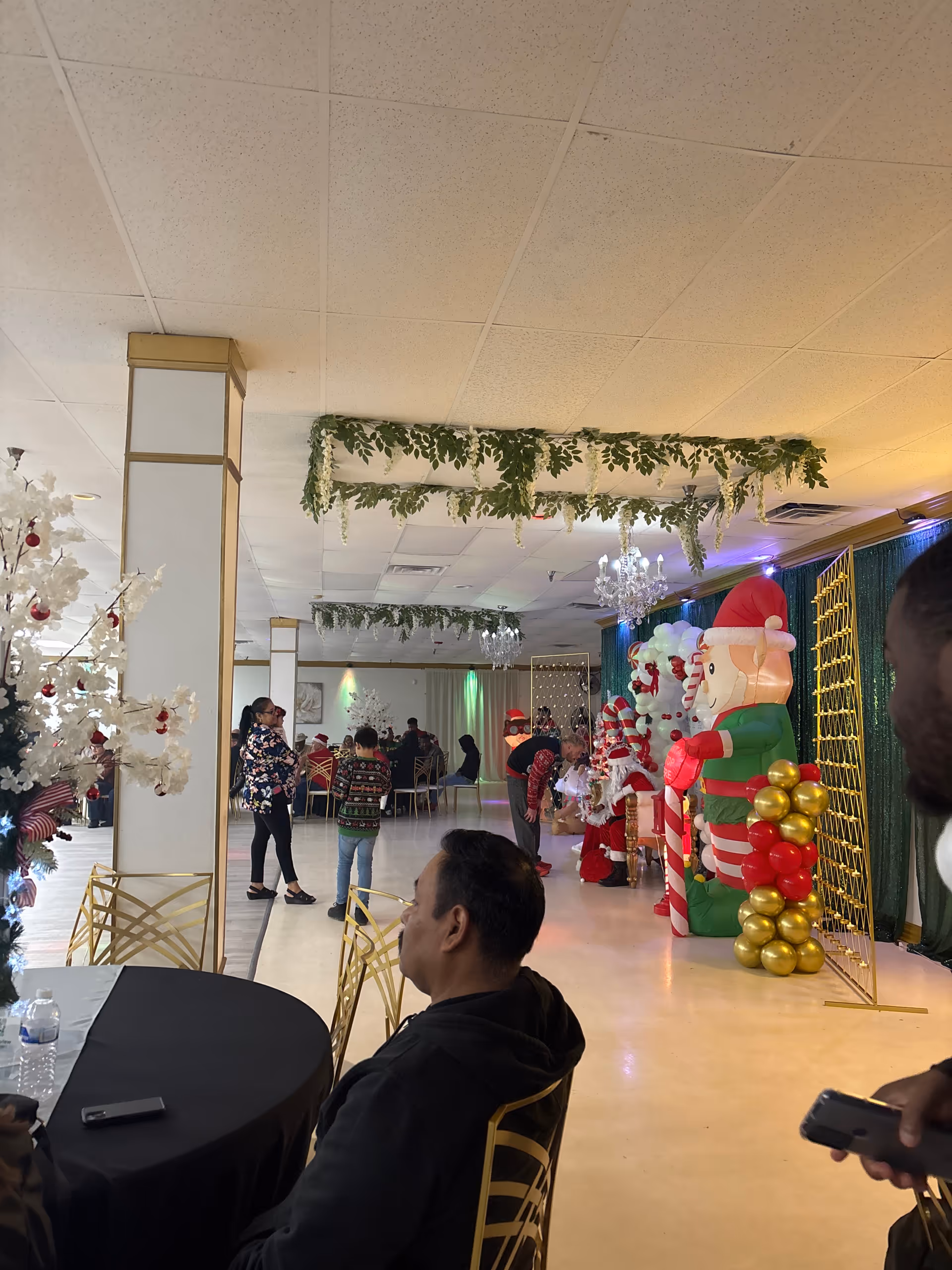 Indoor event hall with Christmas decorations, balloons, inflatable Santa, and people gathered.