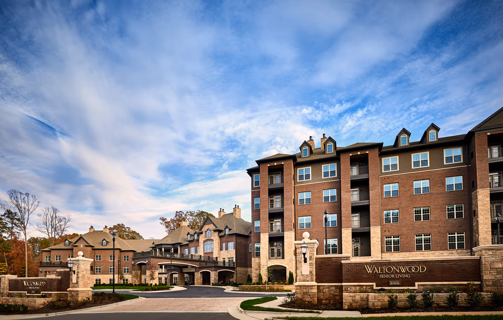 A large brick multi-story senior living building (Waltonwood) with an arched entrance and landscaped driveway under a blue sky.