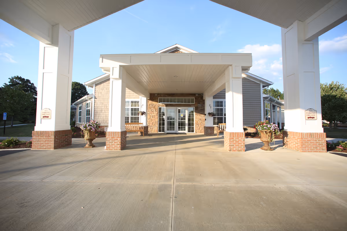 Entrance of a senior living facility with a covered driveway supported by white pillars with brick bases. There are flower pots and benches near the entrance doors, and the building exterior features stone and siding. The sky is clear with a few clouds.