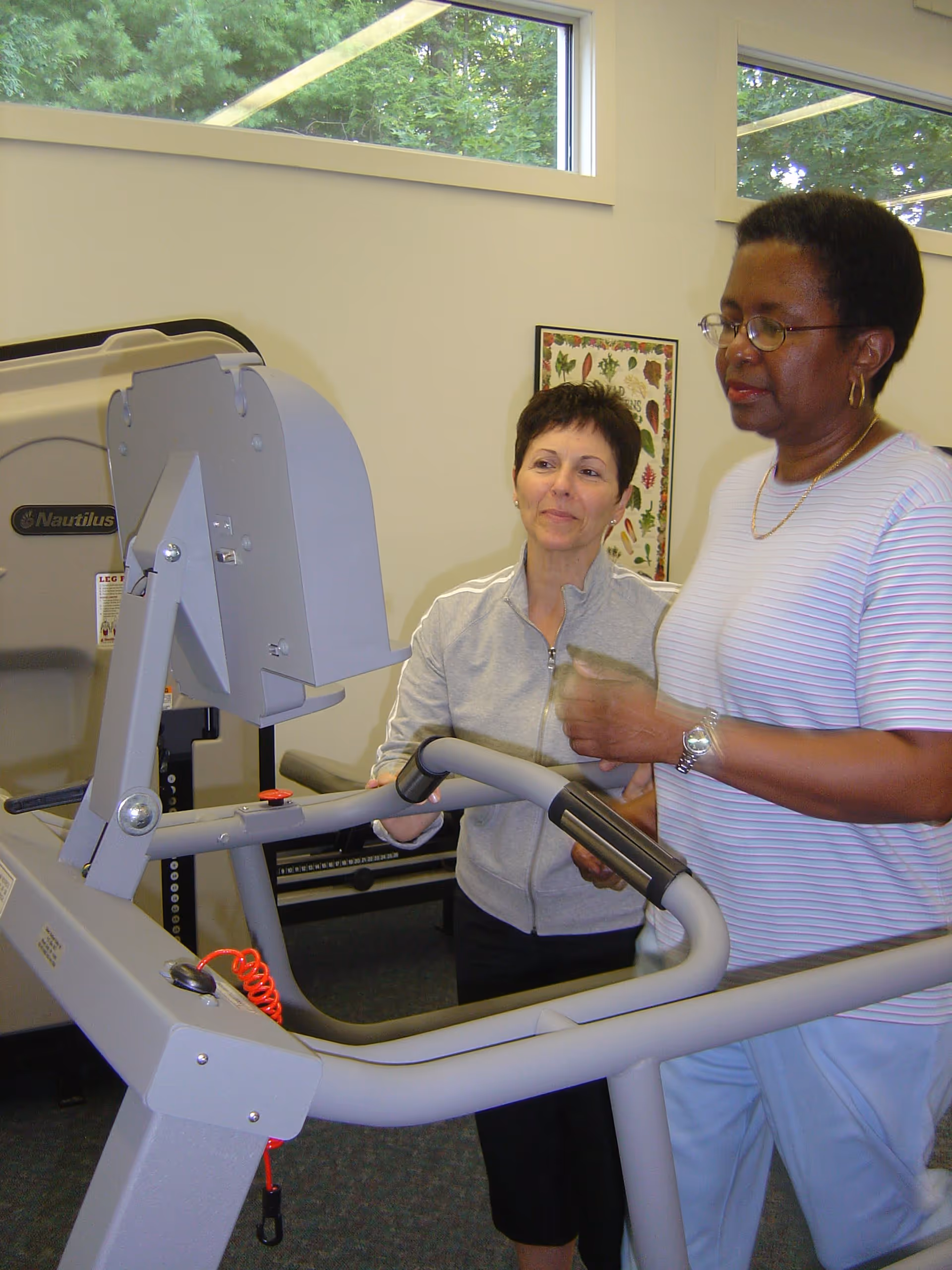 Two women in a fitness room at McLean a CCRC, one woman is using a treadmill while the other stands beside her, possibly providing assistance or instruction. The room has large windows with a view of green trees outside and exercise equipment in the background.