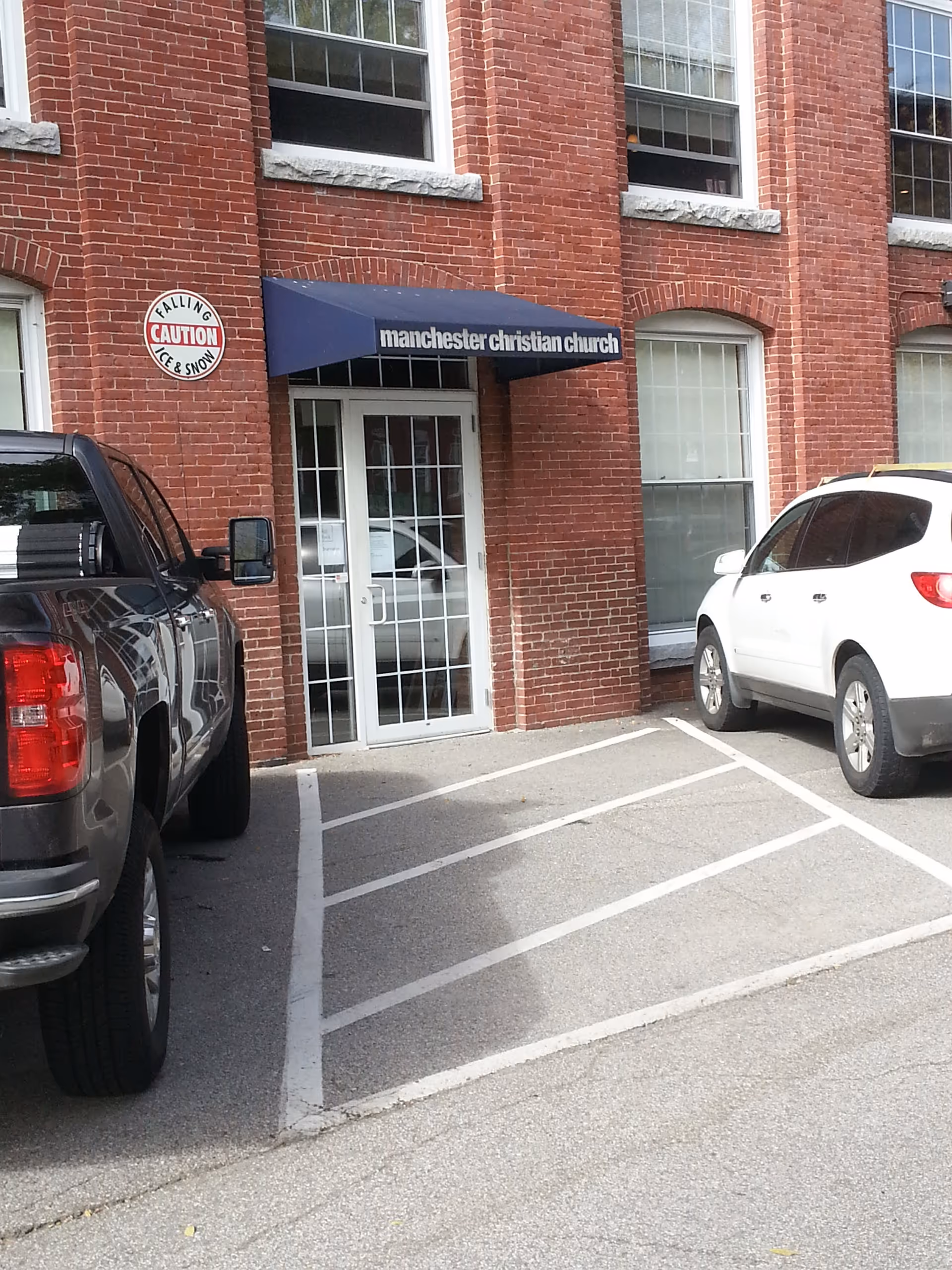 Brick building with blue awning and entrance reading Manchester Christian Church, with parked cars outside.