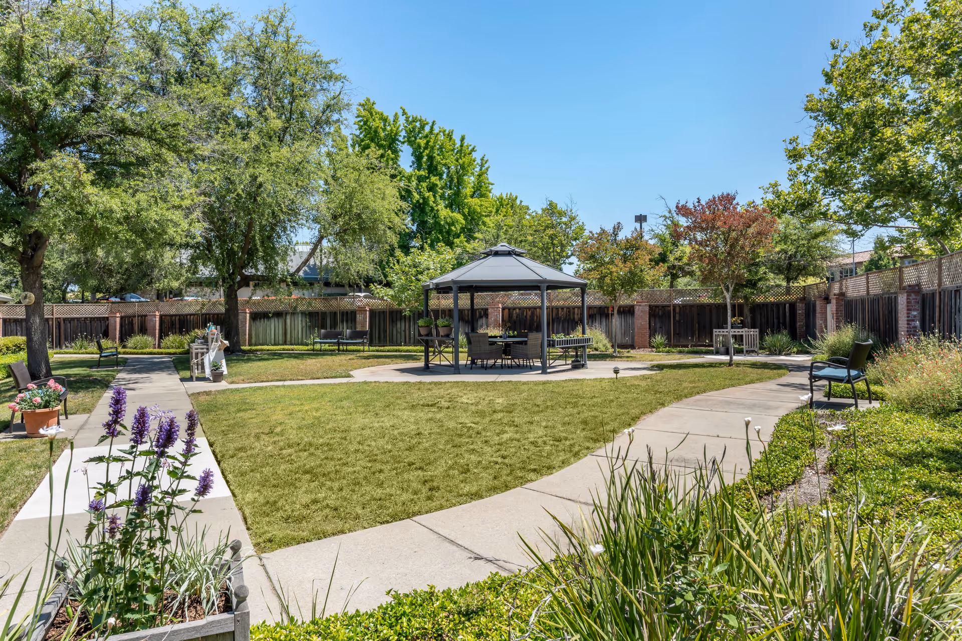 A well-maintained outdoor garden area with a paved walkway curving around a grassy lawn. There is a gazebo with seating in the center, surrounded by trees, plants, and benches along the path. The sky is clear and blue.
