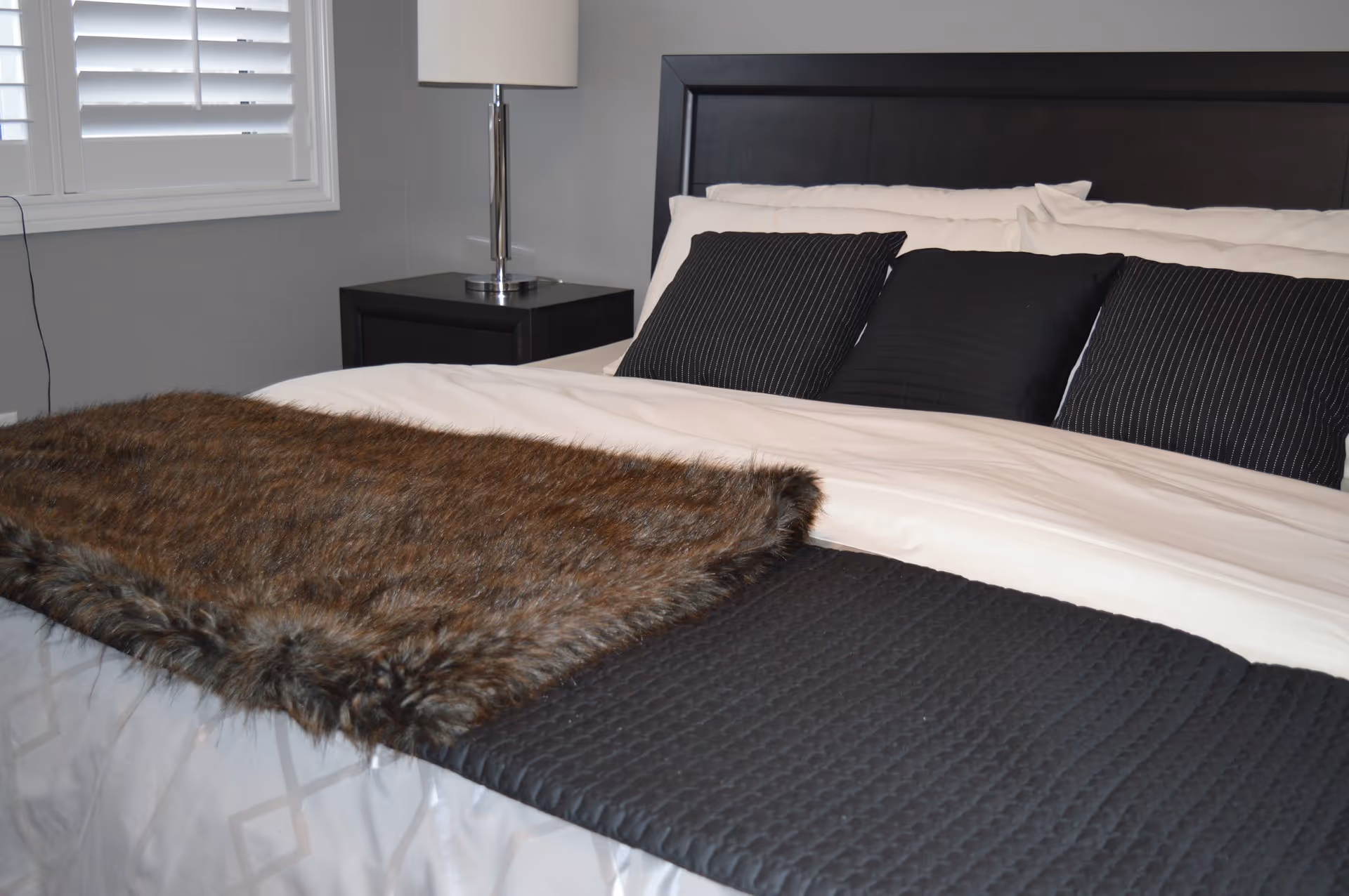A neatly made bedroom with a dark headboard, white bedding, black pillows, and a fur throw.