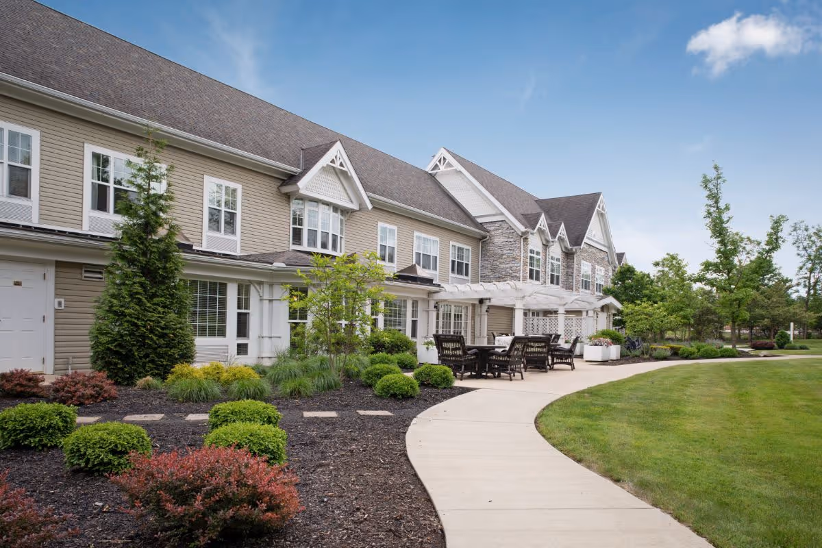 Exterior view of a two-story senior living building with a paved walkway, patio seating, and landscaped lawn and shrubs.