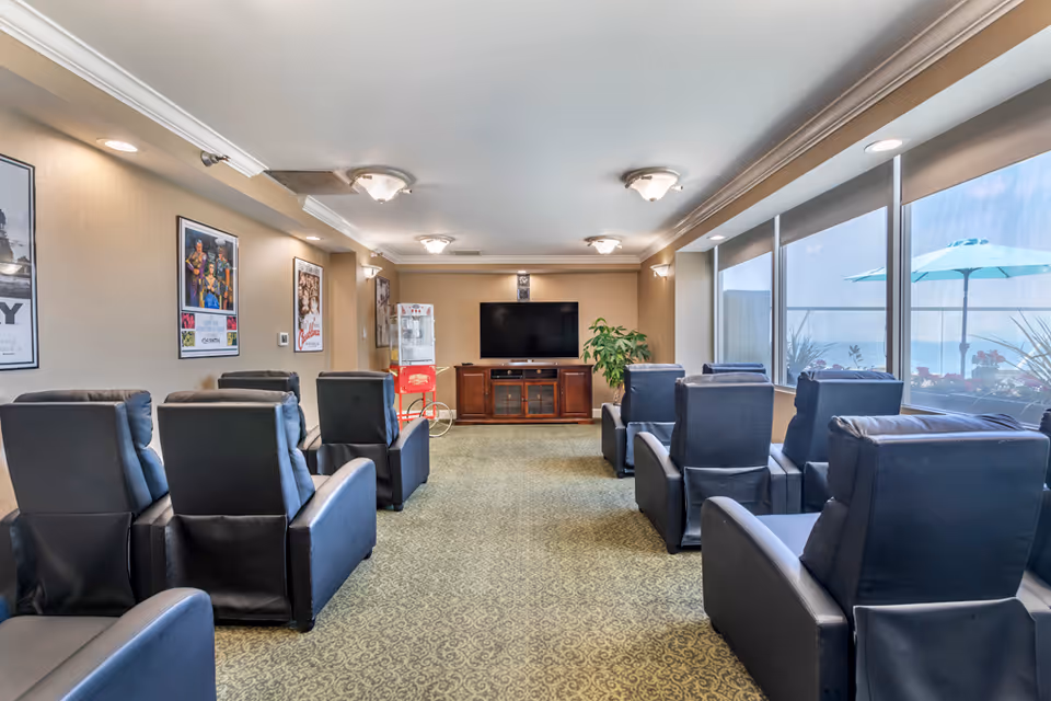 A spacious senior living lounge with multiple recliner chairs, a TV, and large windows.