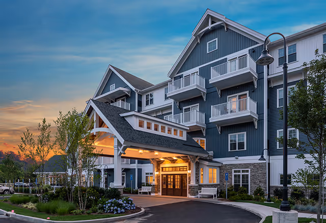 Front entrance of a multi-story senior living building with a covered porte-cochere, balconies, landscaped beds, and a lit evening sky.