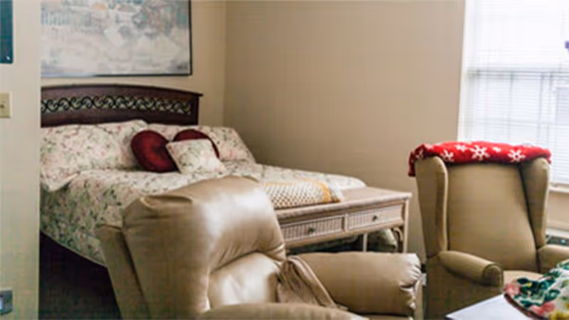 A bedroom with a floral bed, decorative pillows, and two recliner chairs near a window.
