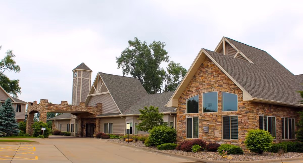 Exterior view of a senior living facility building with stone and siding facade, large windows, a clock tower, and a covered entrance. The surrounding area includes landscaping with bushes and trees, and a paved driveway.