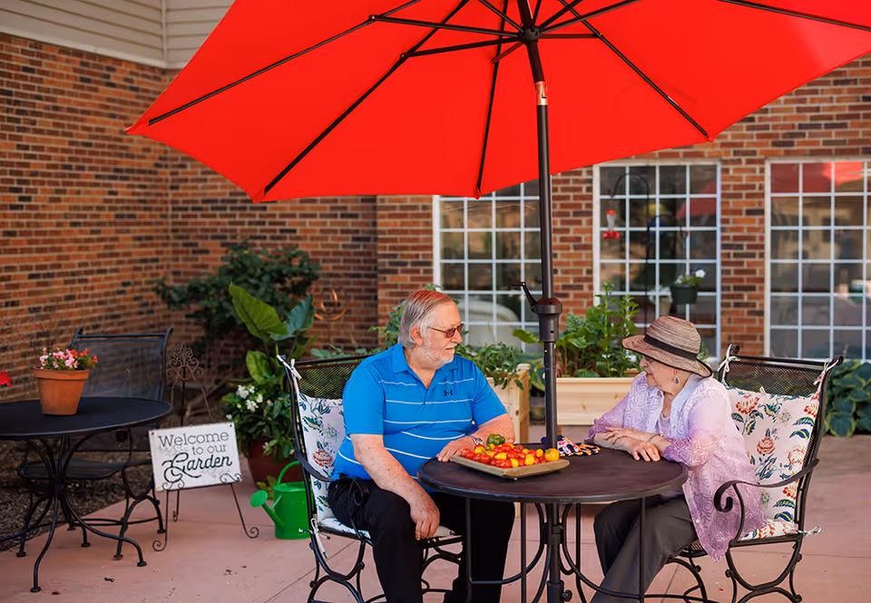 An elderly man and woman sitting at a round outdoor table under a large red umbrella, engaged in conversation. The table has a tray with colorful cherry tomatoes. They are seated on metal chairs with floral cushions. Behind them is a brick building with large windows, potted plants, and a sign that reads 'Welcome to our Garden'.