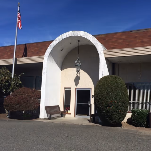 Front entrance of a retirement community building with an arched doorway, bench, trimmed shrub, and an American flag.